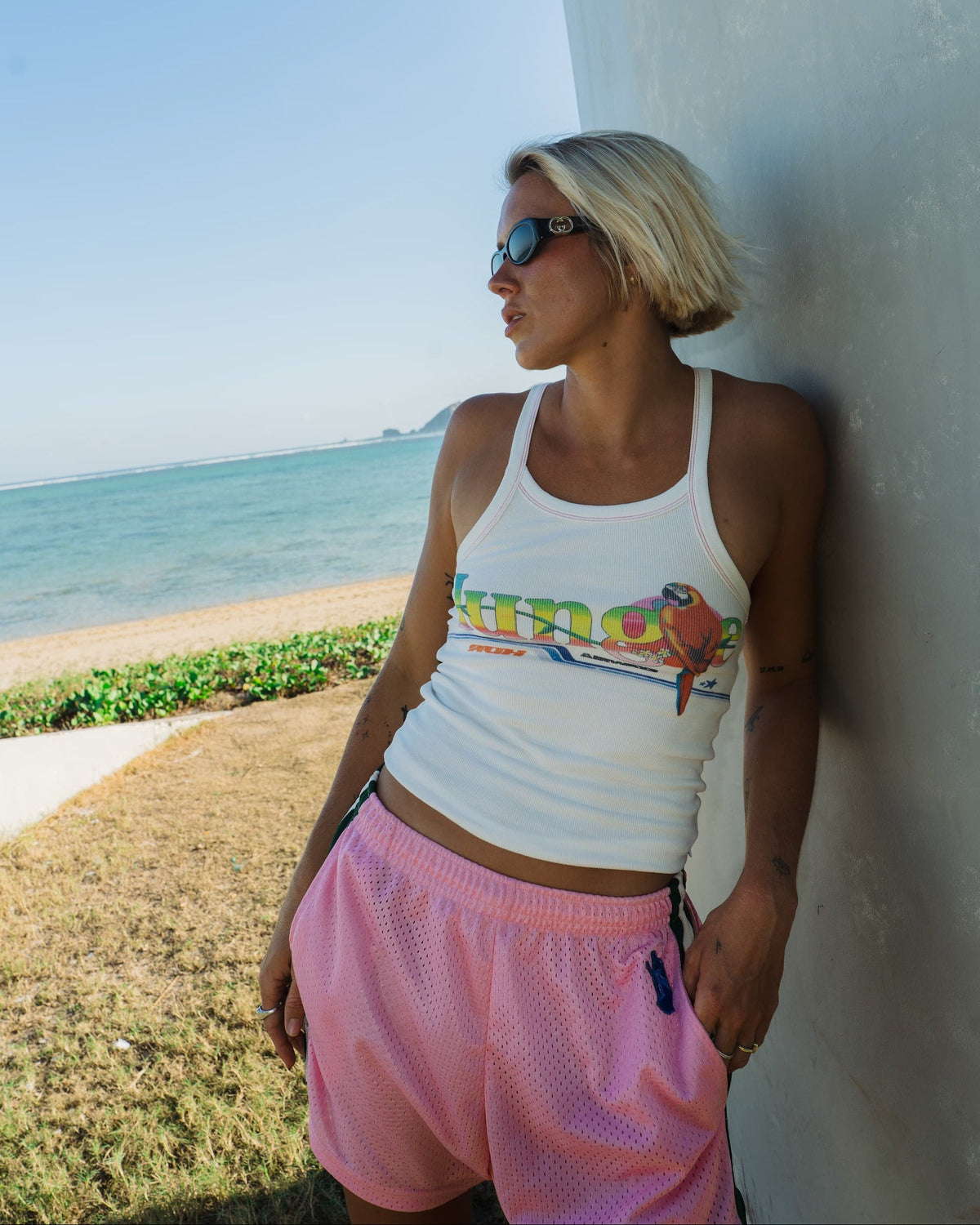 A woman with blonde hair and sunglasses sits on a black ATV in front of a wooden wall, wearing a white tank top with a colorful graphic and pink shorts, carrying a large, vibrant hand-crocheted striped bag.